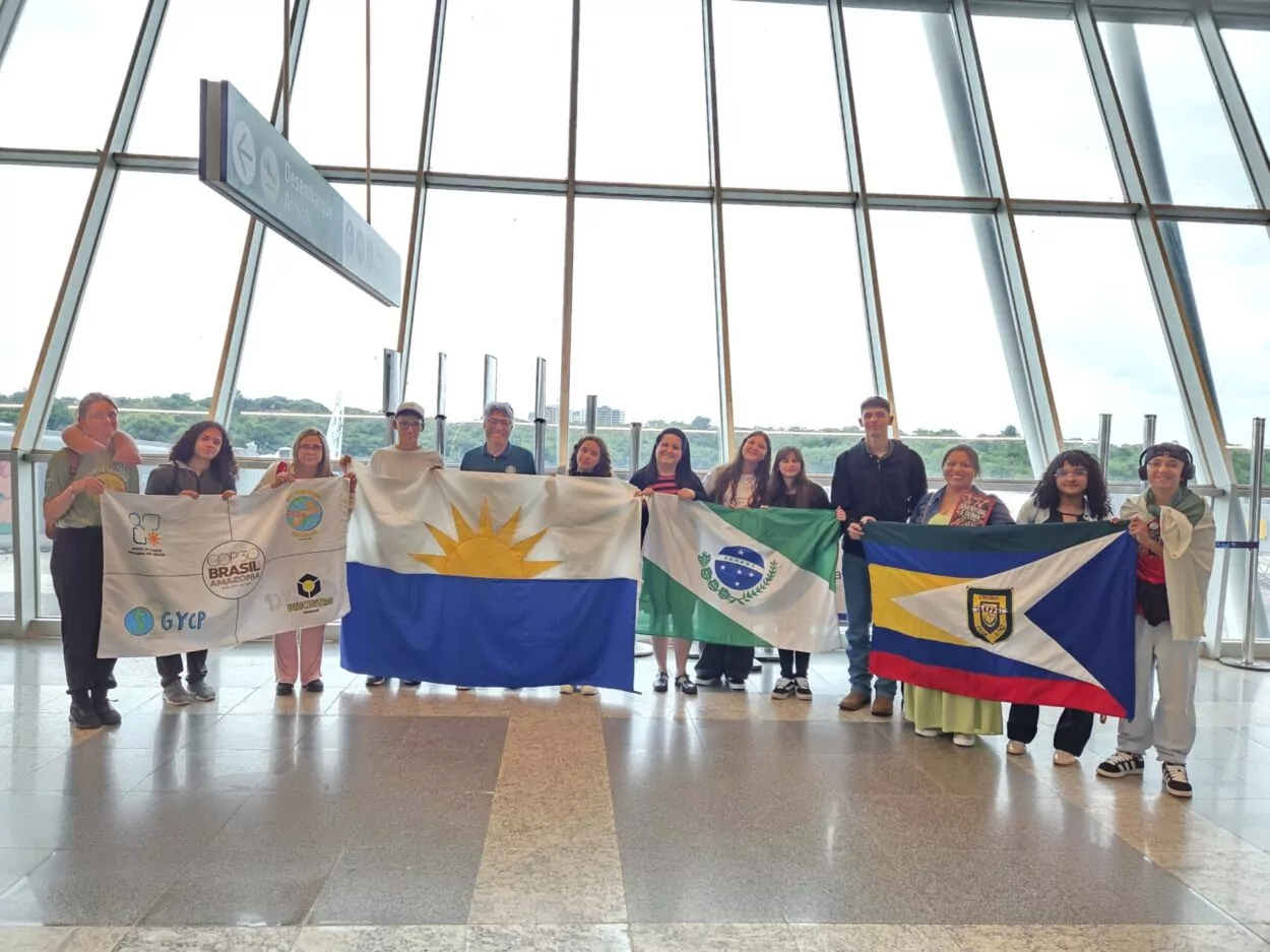 Um grupo de estudantes no saguão de um aeroporto, enfileirados lado a lado, seguram quatro bandeiras no total, uma é do evento COP30 com símbolos da Rede de Clubes Paraná Faz Ciência, do projeto de extensão da Unicentro: Nós Propomos!, do Global Youth Climate Pact (GYCP) - Pacto Mundial de Jovens pelo Clima no Brasil e a logo da Unicentro. Outras bandeiras também são mostradas pelos alunos, como a do Estado do Paraná, da Cidade de Guarapuava e a do Colégio Estadual Padre Chagas, de Guarapuava.