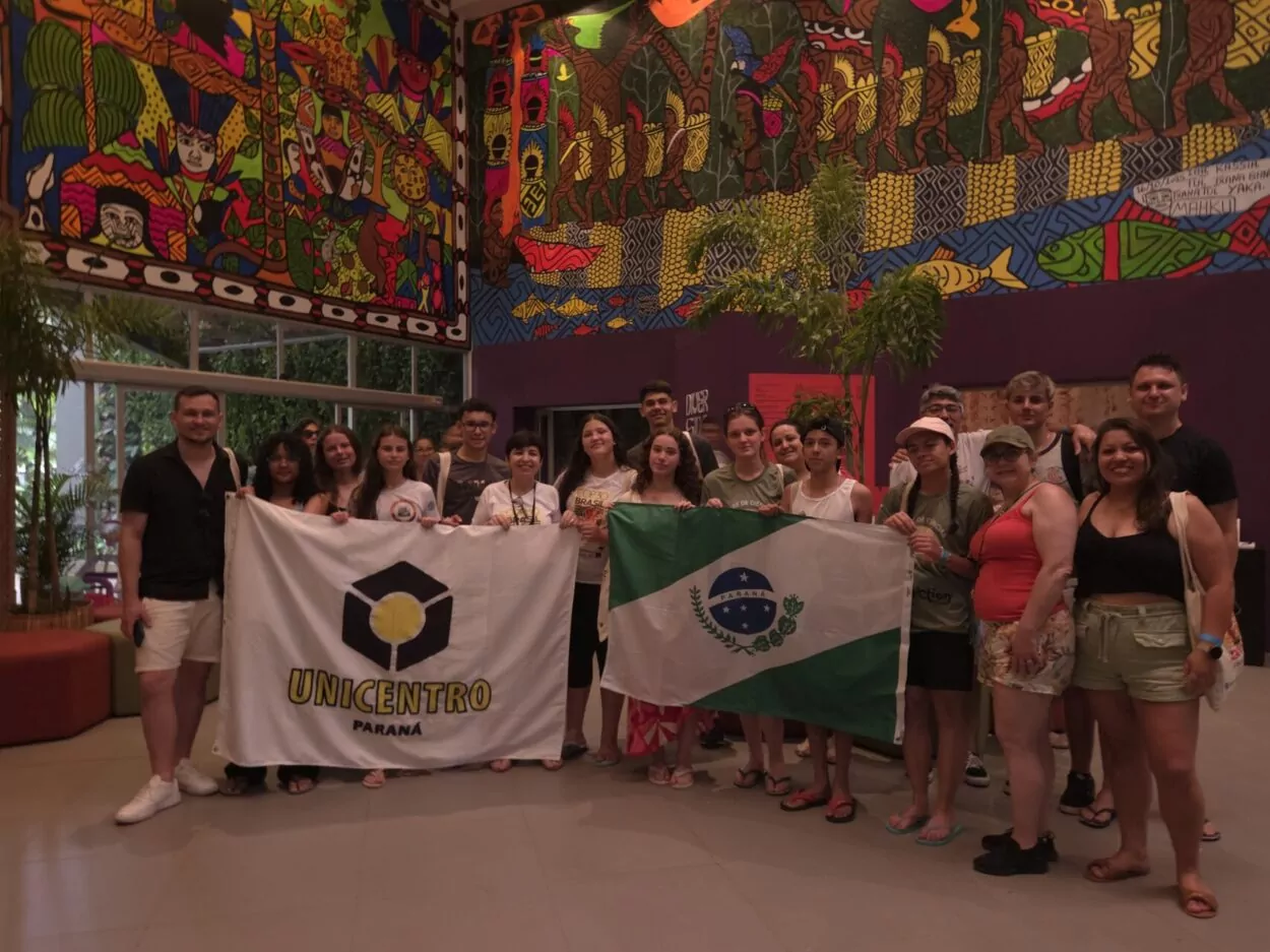 Um grupo de aproximadamente vinte adolescentes e adultos, em pé, posando juntos em um ambiente interno. Eles estão alinhados, segurando duas grandes bandeiras à frente: uma branca com o logo da Unicentro Paraná em preto e amarelo, e outra que é a bandeira do Paraná (verde, branco e azul). O grupo veste roupas casuais de verão, como shorts e camisetas. A cena é emoldurada por uma grande parede ao fundo, totalmente coberta por um mural ricamente colorido, vibrante e detalhado, que apresenta figuras humanas e padrões que remetem à arte folclórica ou indígena, sob uma iluminação interna suave.