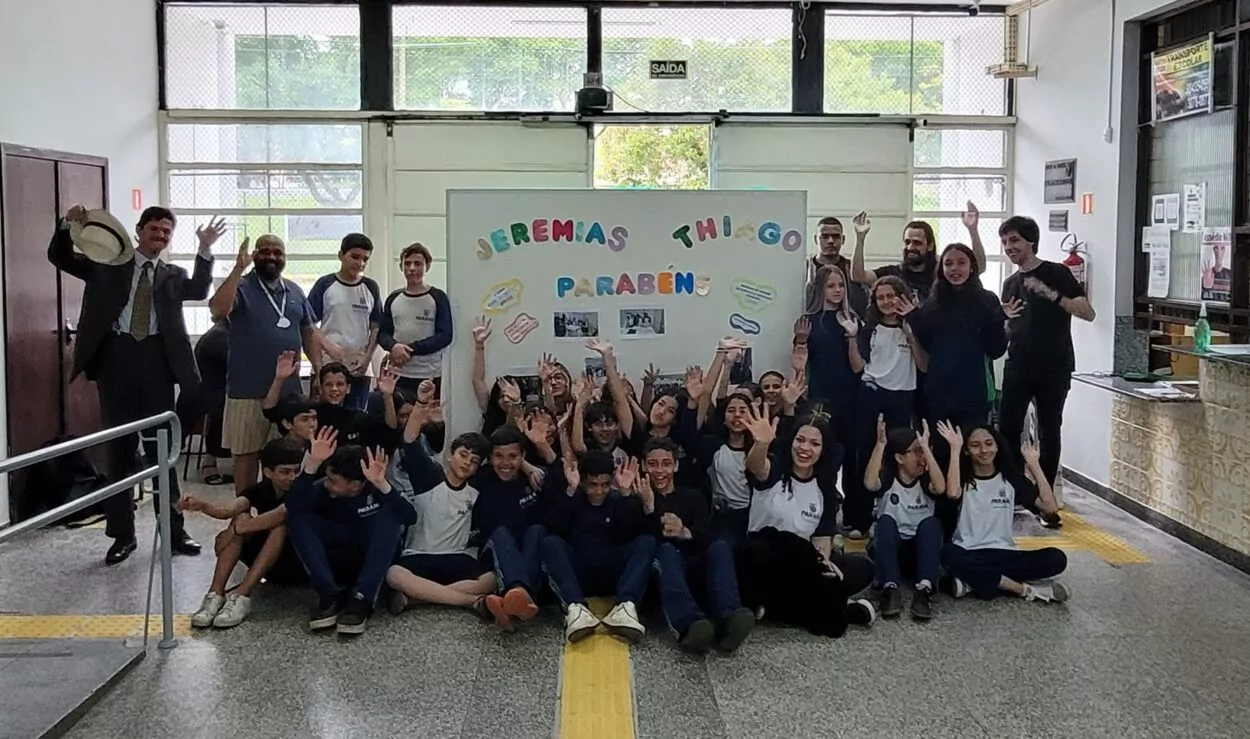 Fotografia em ambiente interno de uma escola. No centro da imagem, há um grande painel branco decorado com letras coloridas formando a frase “Jeremias Thiago Parabéns”, acompanhado de pequenos balões de fala com mensagens e fotos das atividades do clube. Em frente ao painel, sentados no chão, está um grupo numeroso de estudantes, a maioria usando uniforme azul e branco. Atrás deles, outros estudantes estão em pé ao lado do painel. À esquerda, aparece um homem de chapéu claro, camisa social e gravata, e outro homem de camiseta azul. À direita, há mais professores e estudantes em pé. O grupo posa para a foto em clima de celebração, sugerindo um momento de homenagem ou reconhecimento ao trabalho realizado pelo clube na escola.