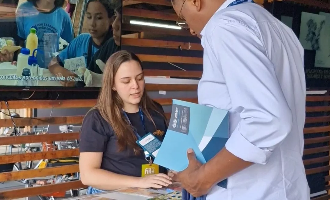 A imagem mostra uma das integrantes da equipe do NAPI PRFC apresentando o projeto para um homem alto. Atrás dela, há uma televisão com um vídeo institucional. No balcão em sua frente, há livros, marca-páginas e a pesquisa de percepção pública desenvolvida pelo NAPI. O homem segura um livro também, sobre espaços de divulgação científica.