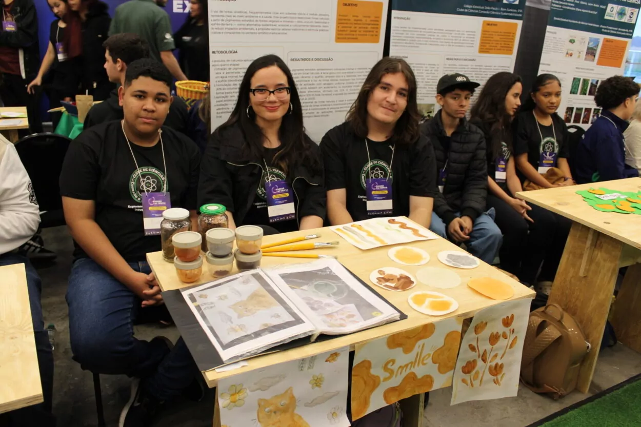 Três estudantes estão sentados atrás de uma mesa de exposição em um evento científico. Eles vestem camisetas pretas com o logotipo de um clube de ciências e crachás roxos no pescoço. À frente deles, a mesa exibe potes de vidro com pigmentos naturais, pequenas amostras de tintas artesanais em tons terrosos, pincéis e folhas com pinturas de gatos, flores e círculos coloridos. A mesa também tem cartazes decorados com desenhos em tons de marrom e laranja, incluindo a palavra “Smile”. Atrás do grupo, há painéis de pesquisa e outros estudantes sentados, indicando que o evento está movimentado.
