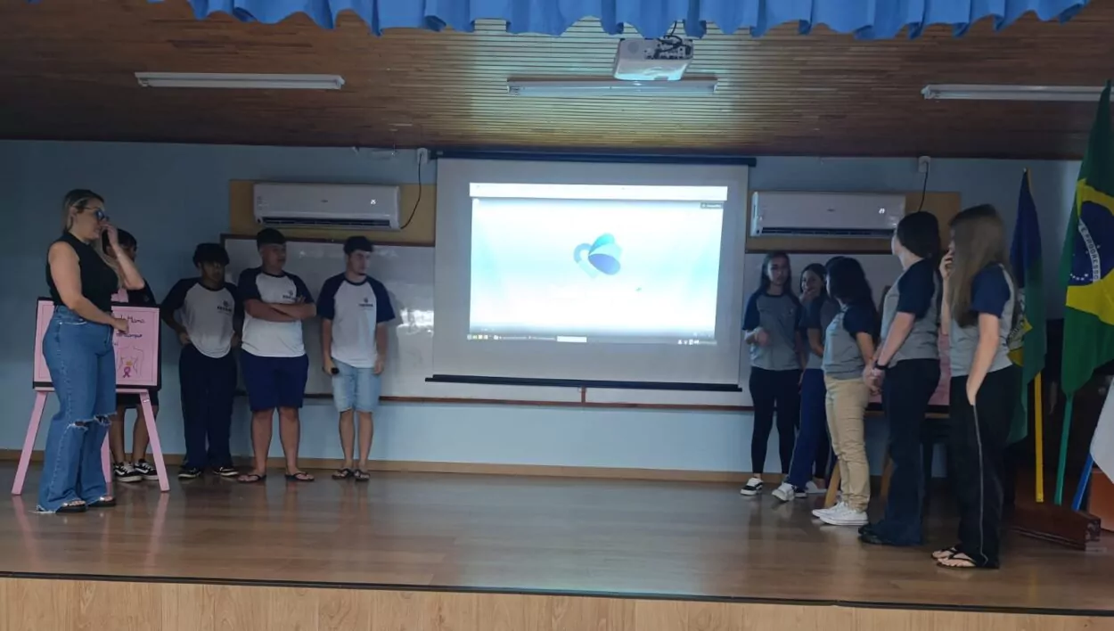 A galeria registra momentos da Culminância do Colégio Estadual Guilherme de Almeida, em Santa Izabel do Oeste (PR). Em um auditório com carteiras enfileiradas, palco, cortinas e projetor, professores e clubistas apresentam os projetos desenvolvidos ao longo do ano de 2025. Há, ainda, imagens de um panfleto intitulado 'Agrotóxicos e o Câncer de Mama' e outros momentos marcantes do clube.