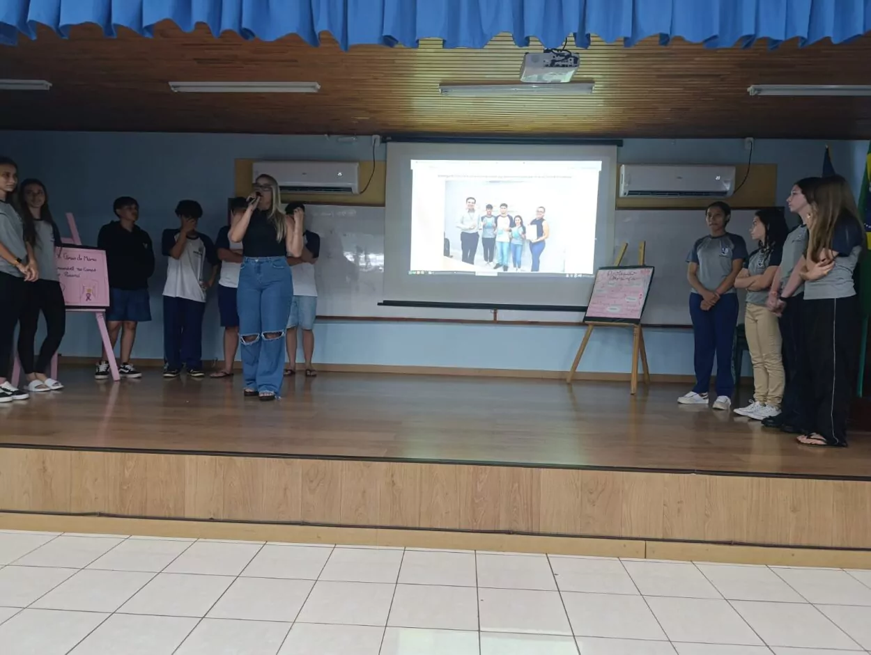 A galeria registra momentos da Culminância do Colégio Estadual Guilherme de Almeida, em Santa Izabel do Oeste (PR). Em um auditório com carteiras enfileiradas, palco, cortinas e projetor, professores e clubistas apresentam os projetos desenvolvidos ao longo do ano de 2025. Há, ainda, imagens de um panfleto intitulado 'Agrotóxicos e o Câncer de Mama' e outros momentos marcantes do clube.