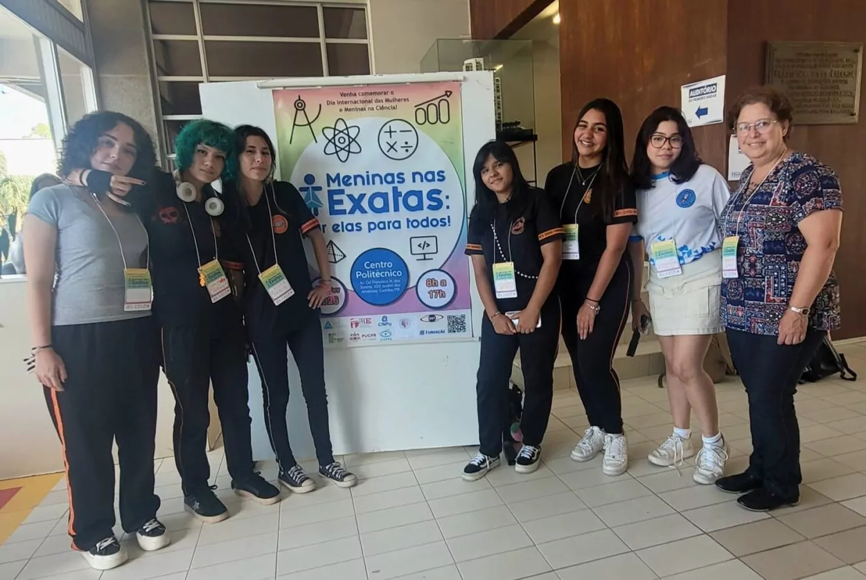 Sete meninas adolescentes e uma professora posam em pé ao lado de um banner do evento “Meninas nas Exatas”, no Campus Politécnico da UFPR. Elas usam crachás e camisetas do clube, em ambiente interno da universidade.