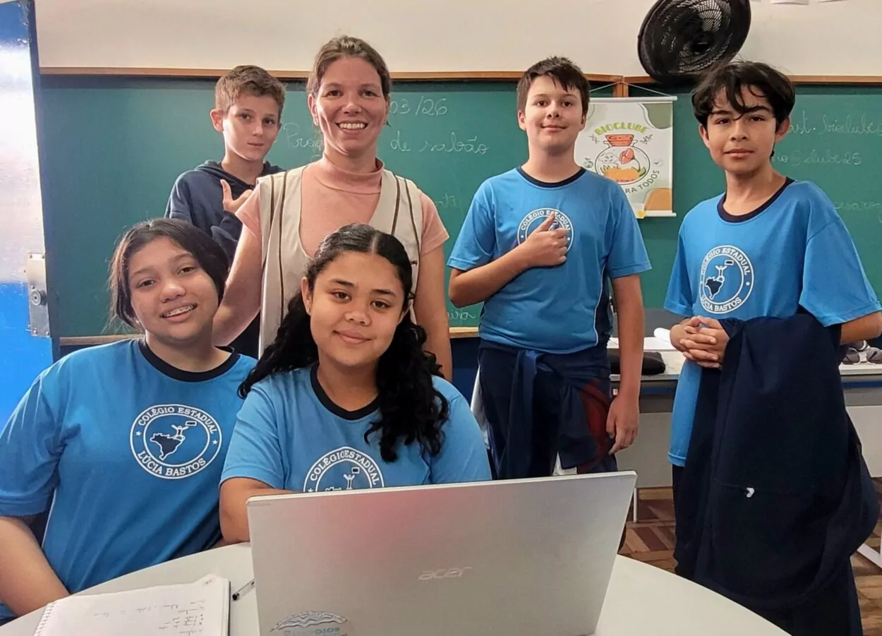 Foto feita dentro de uma sala de aula. Ao centro está uma professora sorrindo, em pé, atrás de uma mesa. À frente dela estão duas estudantes sentadas, uma usando aparelho nos dentes e outra com o cabelo preso em duas tranças, trabalhando em um notebook aberto. Ao redor delas aparecem três estudantes em pé, todos usando camisetas azuis com o símbolo do Colégio Estadual Lúcia Bastos. Ao fundo há um quadro verde com anotações e um cartaz do BioClube. O grupo olha para a câmera enquanto participa das atividades do clube de ciência.