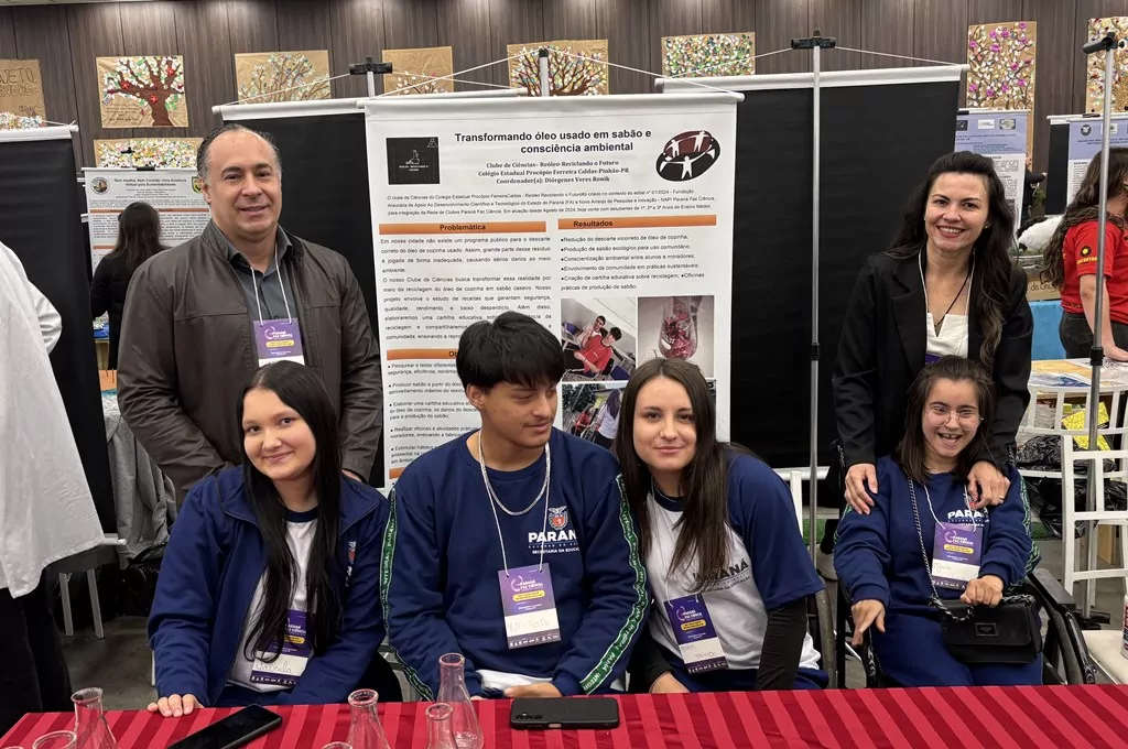 A imagem registra um grupo de seis participantes, entre estudantes e orientador, em uma feira de ciências diante de um banner do projeto "Transformando óleo usado em sabão e consciência ambiental", do Colégio Estadual Procópio Ferreira Caldas (Pinhão-PR). Dois adultos estão em pé nas extremidades, enquanto quatro jovens com uniformes azul-marinho do Governo do Paraná estão sentados à frente de uma mesa com frascos de laboratório, incluindo uma integrante em cadeira de rodas. O painel central detalha a problemática do descarte incorreto de óleo e os resultados obtidos com a produção de sabão ecológico, em um ambiente de exposição repleto de outros estandes e trabalhos educativos ao fundo.