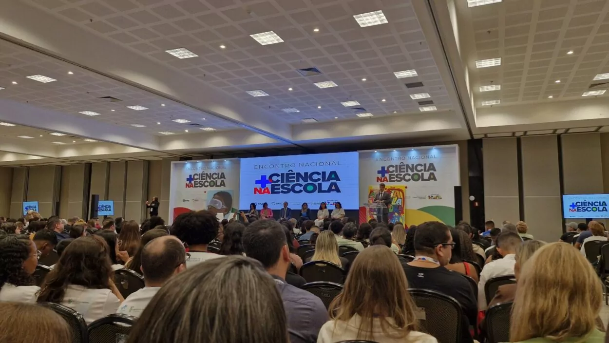 A imagem mostra um auditório lotado durante o Encontro Nacional + Ciência na Escola. As pessoas estão sentadas assistindo a uma apresentação no palco. No palco, há um telão grande com o nome do evento e várias autoridades sentadas, enquanto uma pessoa fala ao púlpito. O ambiente é amplo, bem iluminado e organizado, com foco na programação oficial do evento. A imagem representa um momento formal, provavelmente a abertura ou uma palestra importante.