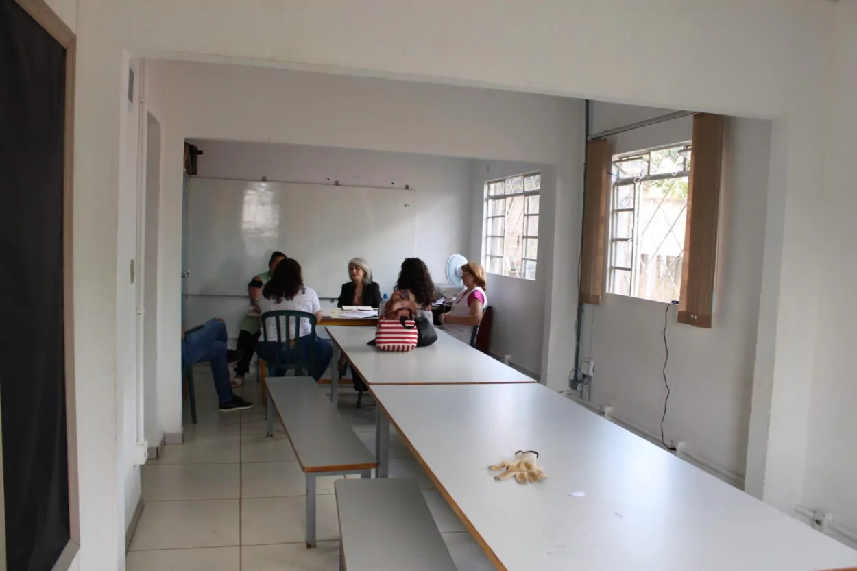 A foto mostra o antigo laboratório. No primeiro plano há mesas longas de madeira com bancos. Ao fundo, um grupo de pessoas está sentado ao redor de uma mesa, conversando e observando alguns papéis. O espaço é iluminado por janelas laterais e possui mobiliário simples, com ventilador e alguns objetos sobre a mesa. 2. Pelo mesmo ângulo da foto anterior, esta foto mostra o novo laboratório de ciências. O espaço possui duas bancadas centrais de mármore com pias e torneiras, além de armários de vidro com vidrarias, equipamentos e modelos didáticos. Ao fundo há uma pequena geladeira e suportes para experimentos, enquanto janelas laterais garantem ventilação e iluminação ao ambiente 3. Em outro ângulo, esta foto também mostra o laboratório novo, desta vez, destaca-se o armário de vidrarias ao fundo, com os equipamentos adquiridos por meio da Rede.