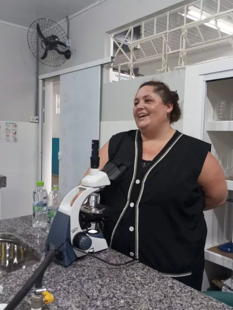 A imagem mostra os clubistas utilizando o novo laboratório de ciências. Eles estão sentados ao redor das bancadas, escrevendo, observando materiais e conversando durante uma atividade. 5. A foto mostra os clubistas realizando uma atividade prática no laboratório. Sobre a bancada estão folhas verdes, recipientes de porcelana, um pilão com pistilo, um béquer com líquido e uma balança digital. Em primeiro plano, uma folha é cortada com uma tesoura e colocada em um recipiente, enquanto outras pessoas ao fundo também manipulam folhas seguindo um roteiro impresso da atividade. 6. A foto mostra um aluno utilizando um microscópio em um laboratório, observando uma lâmina com atenção. O equipamento está sobre uma bancada de pedra, e ao fundo aparecem armários com vidrarias e materiais de laboratório. 7. A foto mostra a professora Rosana ao lado de um microscópio sobre a bancada. Ao fundo aparecem armários com vidrarias e outros materiais científicos, compondo o ambiente de trabalho.
