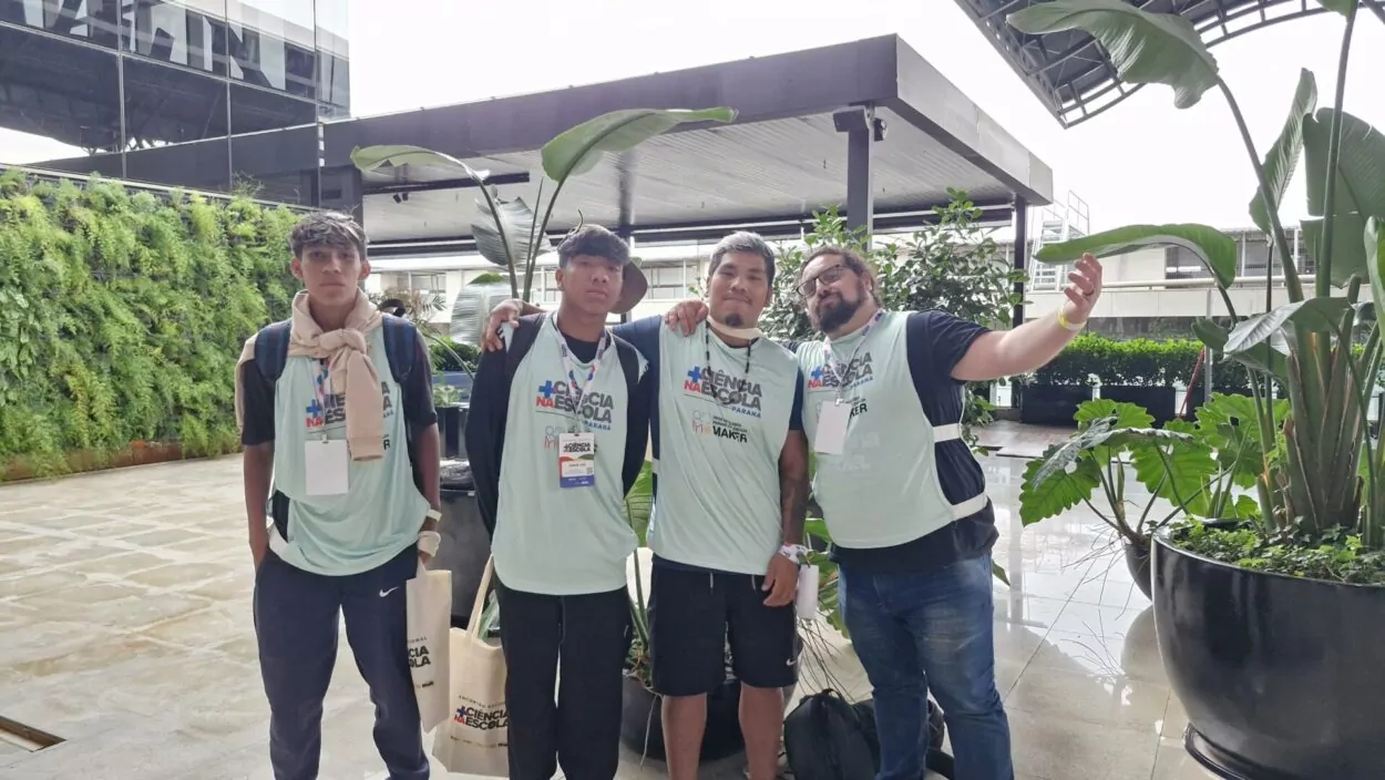 Quatro homens jovens estão lado a lado, posando para uma foto em um espaço moderno com plantas ao redor. Eles usam camisetas claras iguais, com crachás pendurados no pescoço, indicando participação em um evento. Dois deles estão com mochilas. O homem à direita estende o braço, como se estivesse tirando uma selfie ou convidando alguém para a foto. Ao fundo, há um ambiente com cobertura, paredes de vidro e bastante vegetação, sugerindo um local de evento ou instituição.