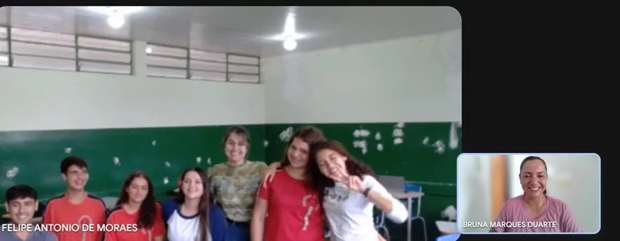 Estudantes reunidos em sala de aula, sentados ao redor de uma mesa com cadernos e materiais escolares. Uma professora aparece em primeiro plano sorrindo, enquanto a bolsista pedagógica Bruna participa por videochamada em destaque na lateral da tela