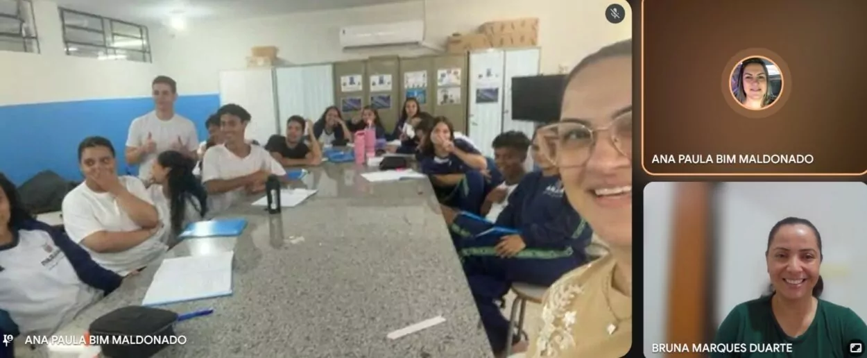 Grupo de estudantes e um professor em sala de aula, organizados em fileiras de carteiras. Um dos alunos segura um caderno aberto. Na tela, a bolsista Bruna aparece em vídeo chamada, acompanhando a atividade