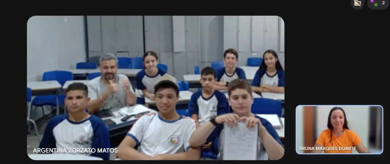 Estudantes reunidos em sala de aula, posando juntos e sorrindo para a câmera. Ao lado, em uma janela de videochamada, a bolsista pedagógica Bruna também aparece sorrindo, indicando participação remota no encontro do clube