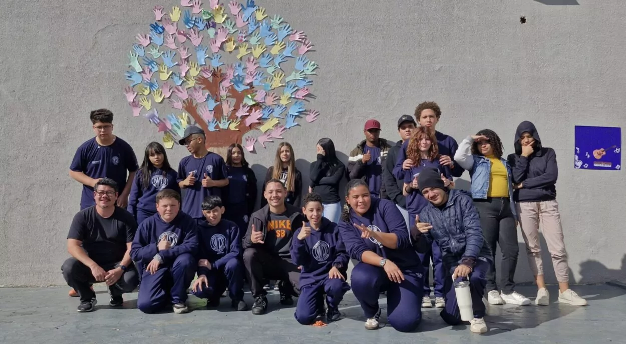 A imagem mostra um grupo grande de adolescentes e alguns adultos reunidos ao ar livre, posando para uma foto. Eles estão organizados em duas fileiras: alguns agachados na frente e outros em pé atrás. A maioria veste uniforme escolar azul-escuro. Ao fundo, há uma parede clara com um desenho de uma árvore feito com vários recortes coloridos em formato de mãos, formando a copa da árvore. À direita, há um pequeno cartaz colorido com desenhos e elementos decorativos. O grupo parece descontraído: muitos estão sorrindo, fazendo gestos com as mãos (como sinal de positivo ou poses informais). A iluminação indica que é dia, com sol forte iluminando a cena.