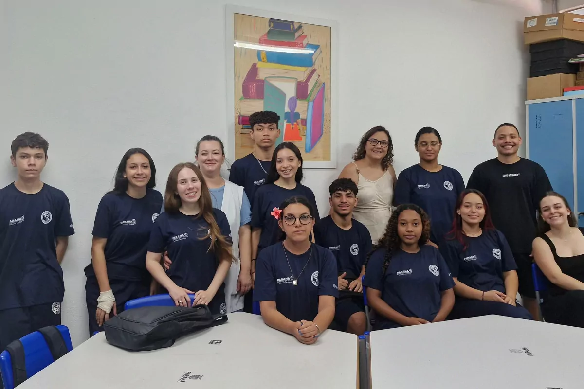 Esta imagem mostra um grupo de estudantes e alguns adultos dentro de uma sala de aula. Eles estão reunidos para uma foto, organizados em duas fileiras: alguns em pé atrás e outros sentados ou apoiados nas mesas à frente. A maioria usa camisetas azul-escuras com um logotipo, sugerindo uniforme escolar. Ao fundo, há uma parede branca com um quadro decorativo que mostra uma pilha de livros coloridos. À direita, aparece um armário azul e algumas caixas empilhadas. As mesas estão à frente do grupo, com mochilas e materiais escolares sobre elas.