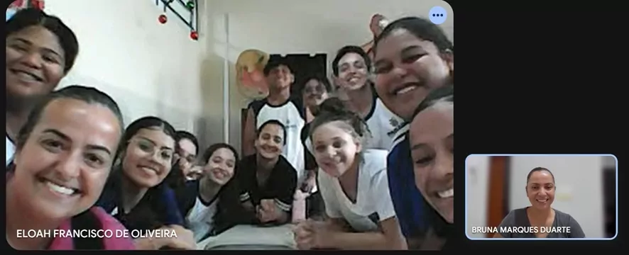 Grupo de estudantes reunidos em uma sala, sorrindo e próximos à câmera em um momento de interação. A imagem parece ser de uma videochamada, com a bolsista pedagógica Bruna visível em um quadro menor no canto direito, também sorrindo.