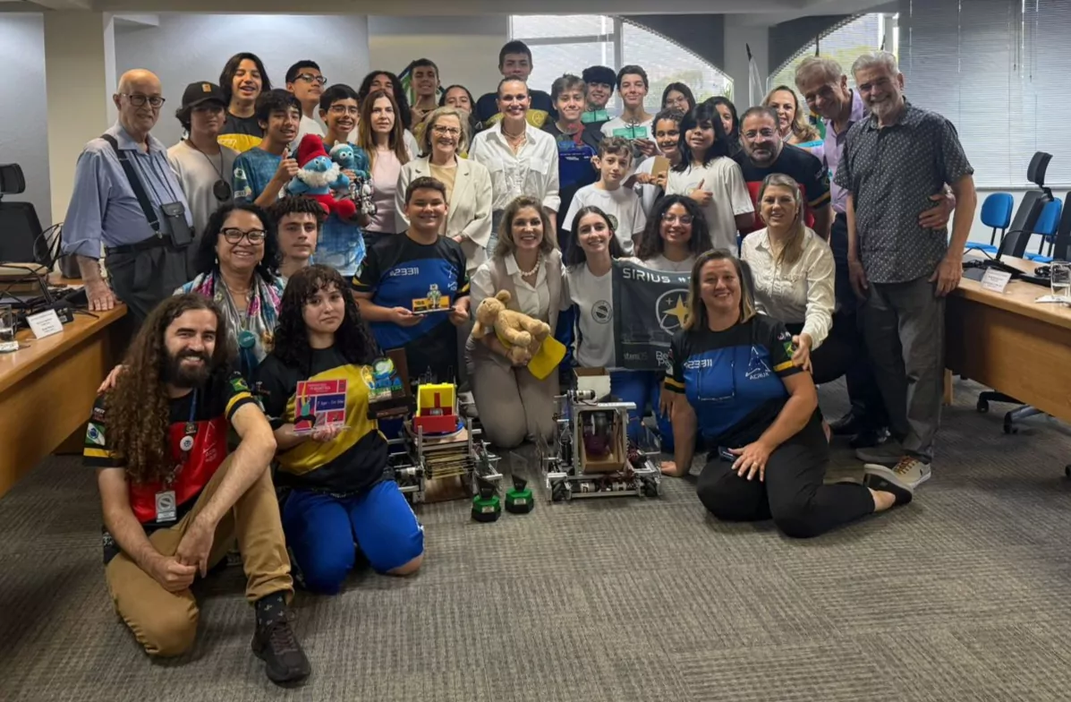 Imagem de um grupo grande de pessoas em um ambiente institucional, posando para foto. Ao centro e ao fundo estão adolescentes e jovens, acompanhados por alguns adultos, formando várias fileiras. A maioria sorri e olha em direção à câmera. Na parte da frente, algumas pessoas estão agachadas ou sentadas no chão, próximas a projetos expostos, incluindo pequenos robôs, maquetes e protótipos tecnológicos. Alguns participantes seguram objetos, como um urso de pelúcia, placas e um banner. Há diversidade de idades, com estudantes e profissionais juntos. Muitos vestem camisetas de projetos ou equipes, algumas com cores escuras e detalhes em amarelo e verde. O espaço possui mesas laterais, cadeiras de escritório e iluminação interna, caracterizando uma sala de reunião ou auditório. A cena transmite um momento coletivo de apresentação e celebração de projetos educacionais e científicos.
