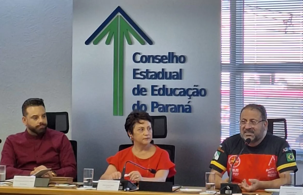 Imagem de uma mesa de reunião institucional. Três pessoas estão sentadas lado a lado, atrás de uma mesa com microfones e copos de água. Ao fundo, há uma parede branca com o logotipo e o nome “Conselho Estadual de Educação do Paraná” em destaque, nas cores azul e verde. À esquerda, um homem de barba, com camisa social vermelha, olha em direção ao centro. No meio, uma mulher de cabelo curto, vestindo blusa vermelha, também está voltada para a direita, com expressão atenta. À direita, um homem de barba e óculos, usando camiseta preta e vermelha com detalhes, fala ao microfone, gesticulando com a mão. O ambiente é formal, com cadeiras de escritório e uma janela com persianas ao fundo.