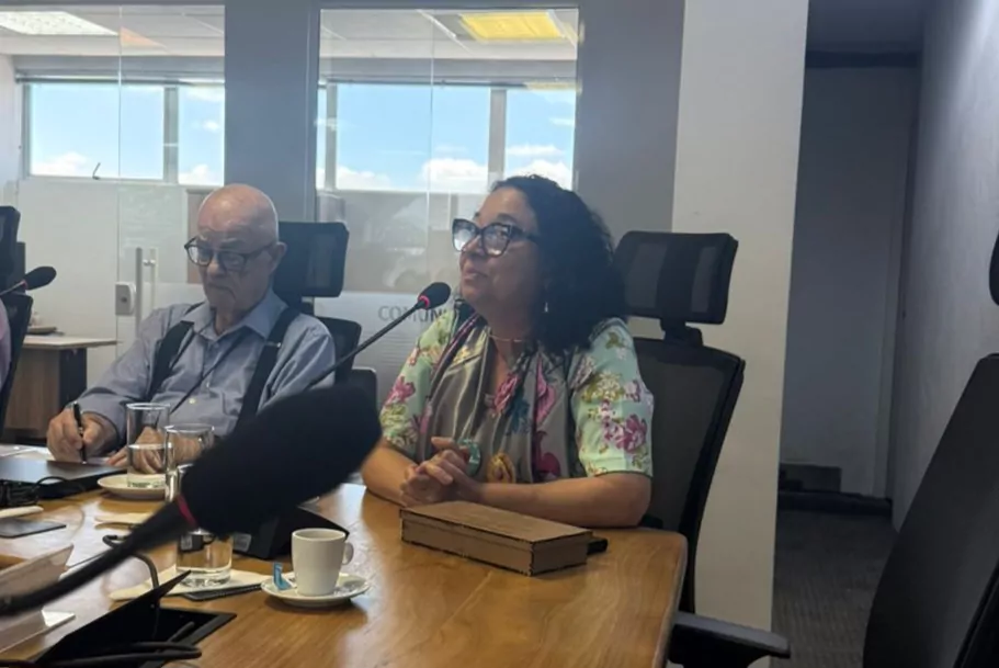 Imagem de uma reunião em ambiente institucional. Em primeiro plano, uma mulher de cabelos cacheados e escuros, usando óculos e uma blusa estampada com flores, fala ao microfone com as mãos apoiadas sobre a mesa. À sua frente, há uma caixa de papelão pequena, uma xícara de café, um pires e um copo de água. À esquerda, um homem idoso, de cabeça calva e usando óculos, camisa clara e suspensórios, está sentado e escreve em um caderno. O ambiente possui uma mesa de madeira, cadeiras de escritório e, ao fundo, uma parede de vidro com janelas que deixam entrar luz natural, mostrando o céu azul do lado de fora. Um microfone aparece em destaque no canto inferior da imagem, indicando que a fala está sendo registrada.