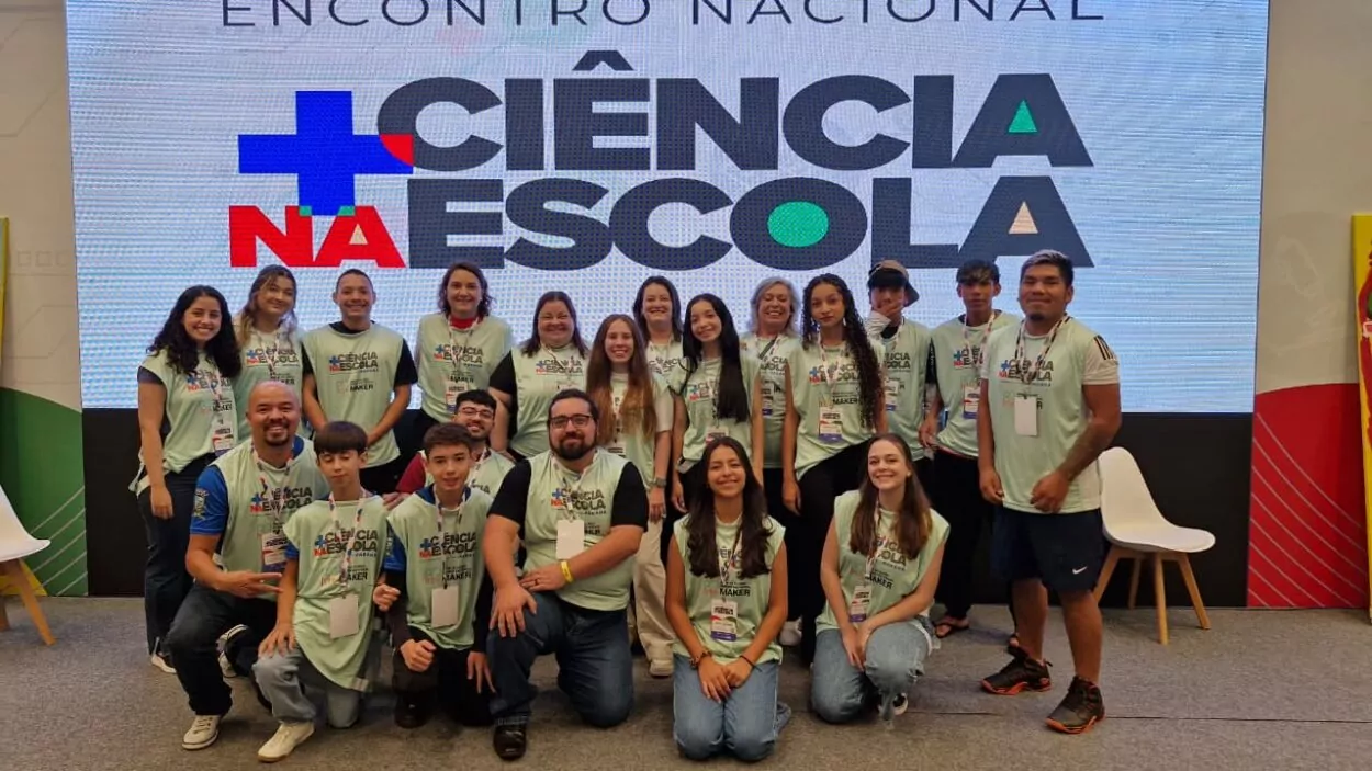 Esta imagem mostra um grupo grande de jovens e adultos posando para uma foto em um evento. Eles usam camisetas verde-claras com crachás, indicando participação oficial. Ao fundo, há um grande painel com o texto “ENCONTRO NACIONAL + CIÊNCIA NA ESCOLA”. O grupo está organizado em duas fileiras (em pé e agachados), sorrindo para a foto. O ambiente é um espaço de evento, com iluminação profissional e estrutura de palco.