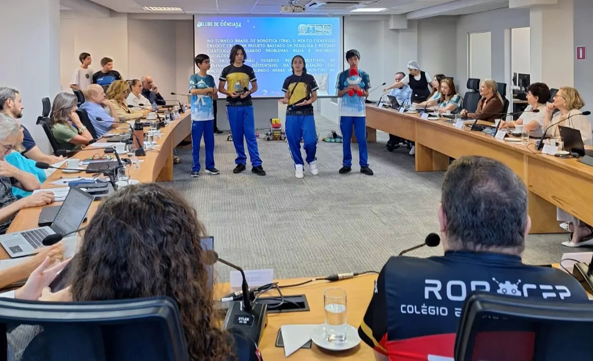 Imagem de uma apresentação em uma sala de reunião institucional. No centro, quatro estudantes adolescentes estão em pé, lado a lado, vestindo camisetas de equipe e calças esportivas, apresentando um projeto. Um deles segura um objeto e outro segura um boneco de pelúcia. Ao redor, em mesas dispostas em formato de “U”, estão diversos adultos sentados, acompanhando a apresentação. Sobre as mesas há microfones, copos de água, laptops e documentos. Ao fundo, um telão exibe um slide com o título “Clube de Ciências” e informações sobre um projeto relacionado à robótica e método científico. O ambiente é amplo, com iluminação interna, cadeiras de escritório e clima formal. A cena mostra um momento de apresentação e avaliação de projetos científicos desenvolvidos por estudantes.