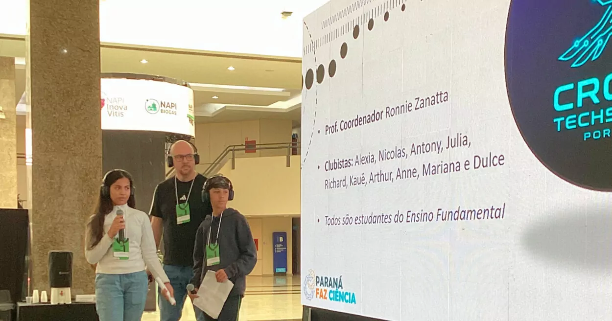 Três pessoas estão em um espaço interno de evento, próximas a um telão de apresentação. Duas estão à frente, utilizando crachás e fones de ouvido, e seguram microfone e papel, como em uma fala ao público. Uma terceira pessoa permanece logo atrás, também com crachá e fones. No telão, aparece um slide com informações do projeto, incluindo o nome do coordenador, lista de participantes e a indicação de que são estudantes do Ensino Fundamental. No canto inferior, há a identidade visual do Paraná Faz Ciência. O ambiente é amplo, com iluminação clara e estrutura de evento ao fundo.