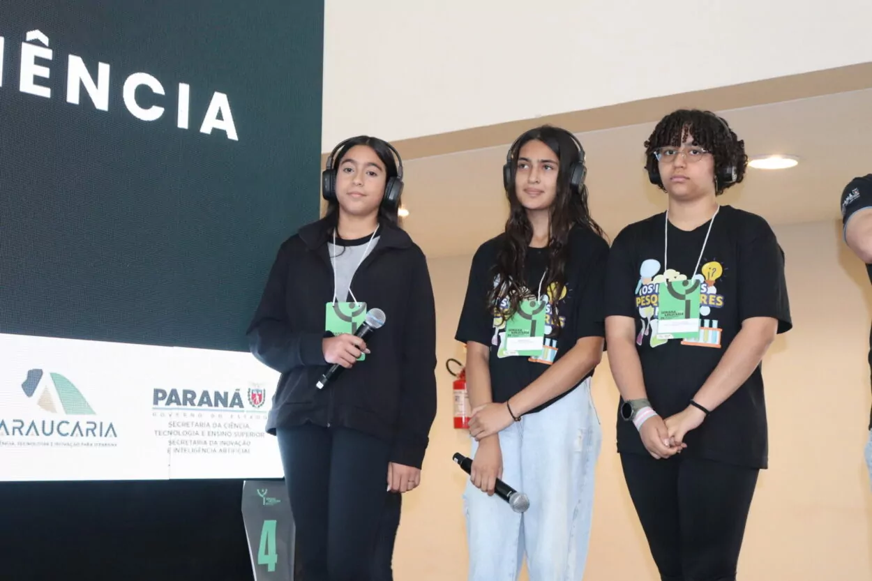 A imagem mostra três adolescentes em um palco durante um evento científico. Elas estão posicionadas lado a lado, voltadas para o público, próximas a um telão de apresentação. Todas utilizam fones de ouvido e crachás de identificação pendurados no pescoço. Duas delas seguram microfones, indicando que estão apresentando ou se preparando para falar. As três vestem roupas escuras, sendo que duas usam camisetas com a identidade visual do grupo “Os Incríveis Pesquisadores Maker”. A postura delas é atenta e concentrada, sugerindo um momento de apresentação formal do projeto.