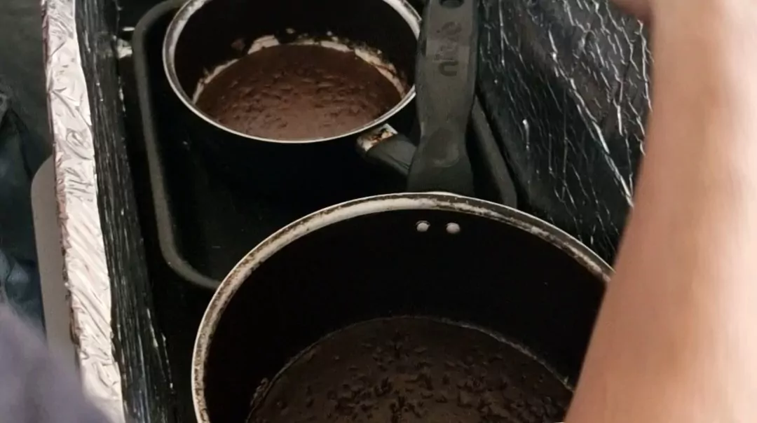 Esta imagem mostra duas panelas pretas vistas de cima, posicionadas lado a lado dentro de uma espécie de caixa ou estrutura revestida com material metálico refletivo (parecido com papel alumínio). Dentro das panelas há bolo de chocolate. Uma delas tem um cabo visível voltado para a direita. Parte do braço de uma pessoa aparece no canto direito da imagem, sugerindo que alguém está manipulando ou observando as panelas. O ambiente parece improvisado, como um experimento ou preparo artesanal.