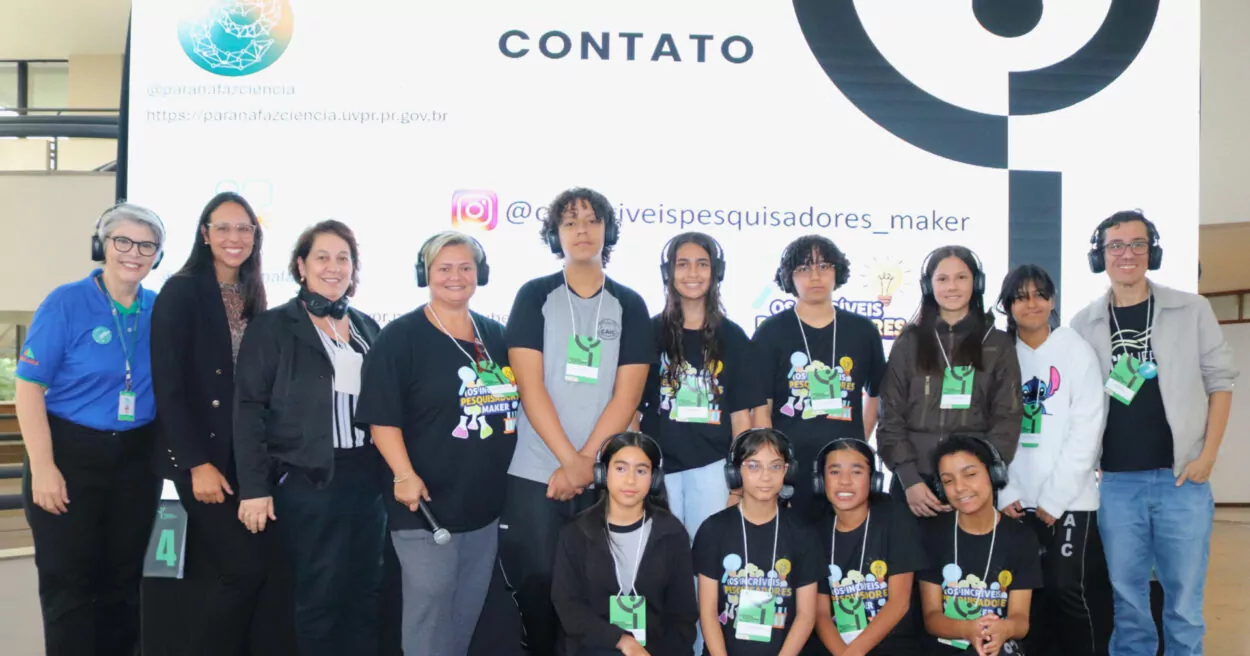 Grupo de pessoas posando para foto em um espaço interno de evento. Ao fundo, há um painel branco com a palavra “CONTATO”, logotipo, endereço de site e um perfil de rede social. As pessoas estão organizadas em duas fileiras: uma em pé e outra agachada à frente. A maioria utiliza camisetas pretas com estampas coloridas relacionadas a ciência e crachás de identificação pendurados no pescoço. Algumas usam fones de ouvido. Há também pessoas com roupas casuais, como jaquetas e camisetas claras. Todas estão voltadas para a câmera, com expressões sorridentes ou neutras. O ambiente é bem iluminado, com estrutura de evento visível nas laterais.
