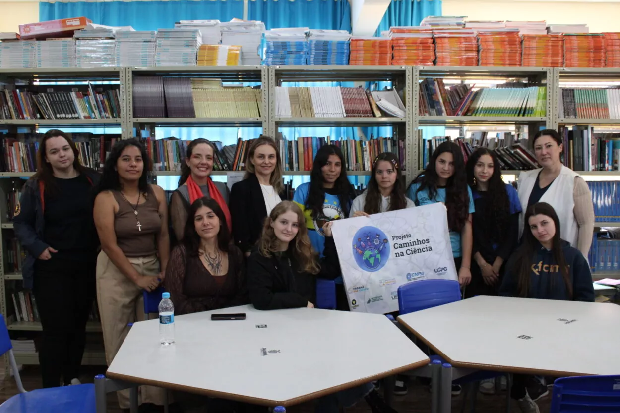 A imagem mostra as alunas e a professora clubista do Lumina, junto as articuladoras e bolsistas do projeto Caminhos na Ciência, elas estão reunidas em uma biblioteca ou sala de leitura. Ao fundo, há estantes cheias de livros organizados por cores e tamanhos. O grupo está posicionado atrás de mesas brancas com cadeiras azuis, algumas pessoas em pé e outras sentadas. No centro, elas seguram um cartaz com o título “Projeto Caminhos na Ciência”, que inclui ilustrações e logotipos institucionais. As pessoas estão próximas umas das outras, olhando em direção à câmera, com expressões neutras ou sorridentes.
