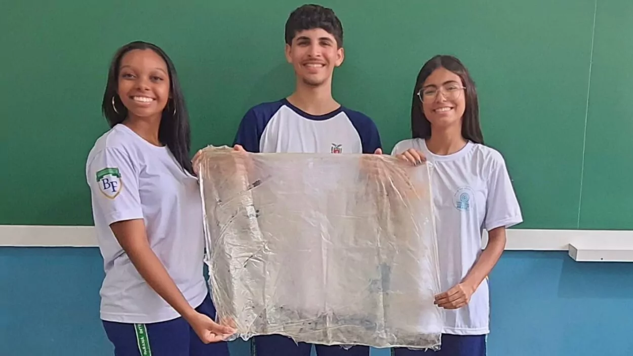 Três jovens estão em pé lado a lado, sorrindo para a câmera, em frente a uma parede com quadro verde (parece uma sala de aula). À esquerda, há uma garota de pele escura, cabelo longo e liso, usando uma camiseta branca de uniforme com um símbolo no braço e calça azul. Ela segura uma das pontas de um grande pedaço de plástico transparente. No centro, há um rapaz de pele clara, cabelo curto e escuro, vestindo uma camiseta branca com mangas escuras. Ele também segura o plástico, ajudando a esticá-lo. À direita, há outra garota de pele clara, com cabelo longo e liso e usando óculos. Ela também veste uma camiseta branca de uniforme e segura a outra ponta do plástico. Os três estão segurando juntos esse grande pedaço de plástico transparente, esticado na frente deles, como se estivessem apresentando um trabalho ou experimento. O ambiente é simples, típico de escola, com parede verde e parte inferior azul.