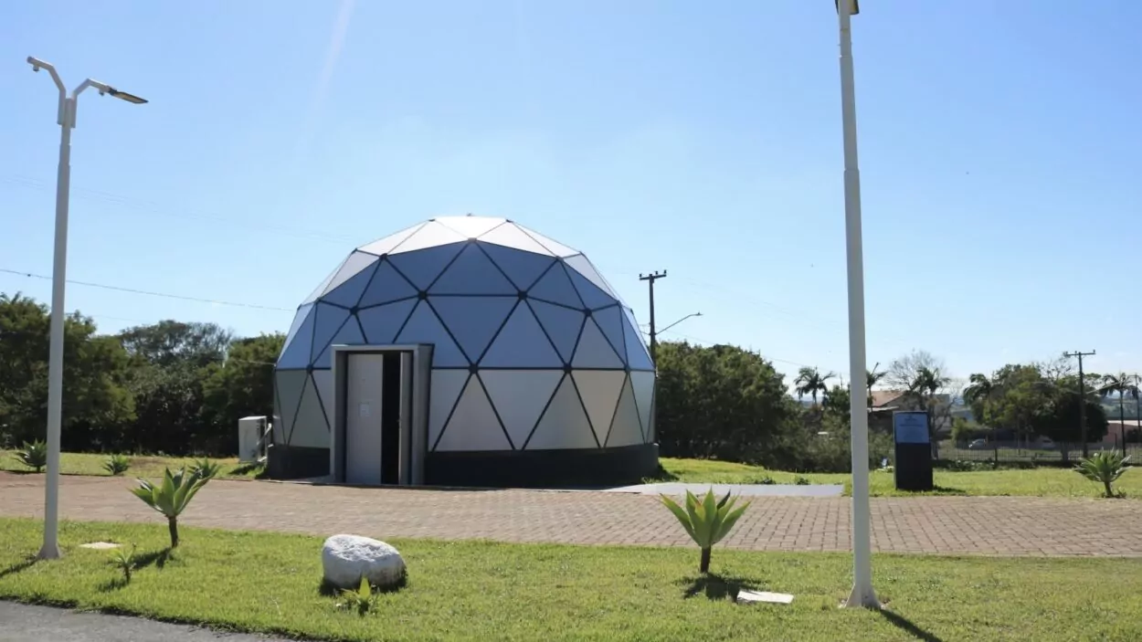 Vista externa do planetário da Universidade Estadual do Norte do Paraná, em Cornélio Procópio. A construção tem formato de cúpula geodésica, com painéis triangulares em tons claros e estrutura escura. A porta de entrada está aberta na parte frontal. O planetário fica em uma área ao ar livre, com gramado bem cuidado, pequenas plantas ornamentais e um caminho de piso intertravado à frente. Há postes de iluminação ao redor e, ao fundo, árvores e algumas construções. O céu está claro e ensolarado, indicando um dia de tempo aberto.