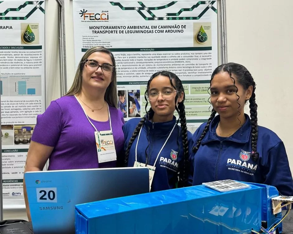 Foto de uma professora e duas estudantes em pé, lado a lado, durante uma feira científica. Ao fundo, há um pôster com o título “Monitoramento Ambiental em Caminhão de Transporte de Leguminosas com Arduino”, apresentado na FECCI 2025. As estudantes usam uniforme azul da rede estadual do Paraná, enquanto a professora veste blusa roxa e crachá de identificação.