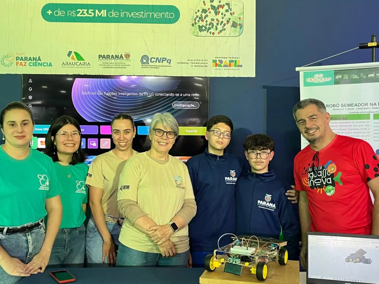 A imagem mostra um grupo de sete pessoas posando em um estande de feira científica e tecnológica. O ambiente parece ser um evento educacional ligado à inovação, pesquisa e robótica. À esquerda, há três mulheres usando camisetas verdes e bege com logos da Rede de Clubes Paraná Faz Ciência,. No centro e à direita, aparecem dois adolescentes usando uniformes azul-marinho da Secretaria da Educação do Paraná. Ambos usam óculos e estão ao lado do projeto apresentado. À direita, há um homem sorridente usando uma camiseta vermelha com a frase “festival iguaçu inova”.Na frente do grupo está um robô de pequeno porte com rodas amarelas, estrutura metálica e de madeira, fios e componentes eletrônicos expostos e sensores e placas eletrônicas aparentes. Ao lado direito da mesa, um notebook exibe na tela um modelo 3D do robô, sugerindo que o projeto foi planejado digitalmente. Atrás do grupo há uma televisão ligada em uma interface da LG Smart TV. Um banner ao fundo menciona o projeto “ROBÔ SEMEADOR NA HORTA ESCOLAR”. Na parte superior aparecem logos de instituições como: Governo do Paraná, CNPq, Governo Federal do Brasil e Fundação Araucária. Também há um destaque escrito “+ de R$ 23,5 MI de investimento”, relacionado ao incentivo à ciência e inovação. A foto transmite um clima de colaboração acadêmica, tecnologia educacional e apresentação de projeto científico escolar.