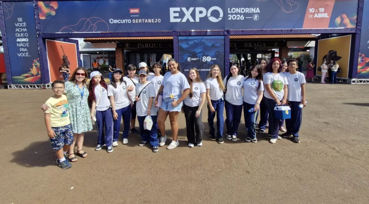 A imagem mostra um grupo de alunos e professores posicionado na entrada de um evento, com um grande portal exibindo “EXPO Londrina 2026”. Os estudantes vestem uniformes e estão organizados lado a lado para uma foto coletiva. Algumas pessoas seguram objetos como garrafas de água e caixas térmicas. O ambiente é externo, com bastante luz natural, indicando um momento de visita ou passeio educativo.