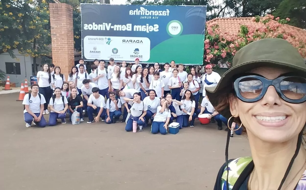 A imagem mostra uma foto em estilo “selfie”, onde uma mulher está sorrindo em primeiro plano, usando chapéu e óculos escuros. Ao fundo, há um grande grupo de estudantes uniformizados reunidos em frente a um banner com o nome “Fazendinha” e “Sejam Bem-Vindos”. O clima é de excursão escolar, com todos posando de forma descontraída ao ar livre.