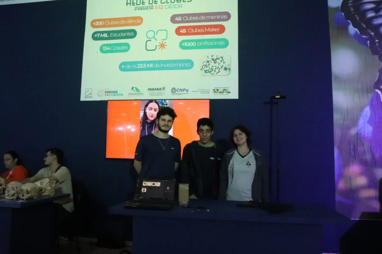 A imagem mostra três jovens posando em um estande de uma feira de ciência e tecnologia. No centro estão dois rapazes e uma moça, em pé atrás de uma mesa de exposição. Sobre a mesa há um notebook aberto e pequenos objetos espalhados, relacionados ao projeto apresentado.Atrás do grupo há uma grande tela exibindo informações do programa “Rede de Clubes Paraná Faz Ciência”. Em uma tela menor ao fundo aparece a imagem de uma jovem falando em vídeo.