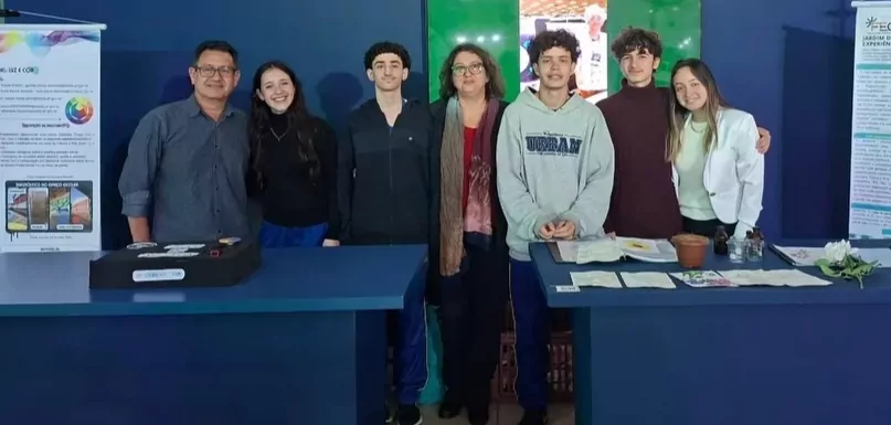 A imagem mostra um grupo de sete pessoas posando em um espaço de exposição científica, durante uma feira de ciências. O grupo é formado por adultos e jovens estudantes. Todos estão lado a lado, sorrindo para a câmera, atrás de duas mesas de apresentação. No centro há uma mulher usando óculos e um cachecol, aparentando ser professora e orientadora do grupo. Há dois banners científicos posicionados nas laterais: à esquerda, um banner sobre um projeto relacionado ao uso de cores e pigmentos. À direita, outro banner com textos científicos, imagens e gráficos sobre experiências interdisciplinares envolvendo artes e ciências. Sobre as mesas há documentos e folhas impressas, recipientes e objetos de experimento, pequenos materiais de demonstração, uma planta/folhagem exposta, e uma caixa de apresentação.