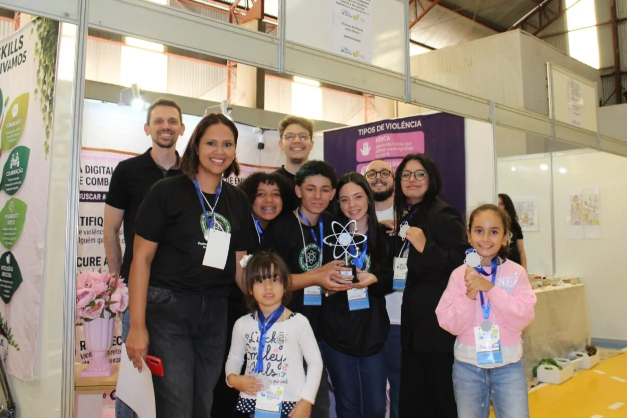 A imagem mostra um grupo de dez adultos, adolescentes e duas crianças posando para uma foto em um estande de feira ou exposição educacional. Todos estão sorrindo e voltados para a câmera, transmitindo um clima de celebração e conquista. No centro do grupo, alguns jovens vestem camisetas pretas com um logotipo científico e usam crachás de identificação pendurados no pescoço. Eles seguram um troféu em formato de átomo, uma premiação de projeto relacionado à ciência, tecnologia e educação. Ao lado deles, há adultos que aparentam ser orientadores ou professores. À frente, duas meninas menores também usam medalhas e crachás. Uma veste blusa branca com estampas coloridas, e a outra usa um moletom rosa claro e segura uma medalha com as duas mãos. O estande ao fundo apresenta painéis informativos. Em um deles lê-se “TIPOS DE VIOLÊNCIA”, com categorias como violência física e psicológica. Outro painel contém textos e elementos gráficos relacionados a habilidades e educação. O ambiente é um pavilhão amplo, com estrutura metálica aparente e iluminação natural entrando pelas laterais superiores. A fotografia registra um momento de confraternização de uma equipe em um evento educacional e científico, destacando o orgulho dos participantes por sua apresentação ou reconhecimento recebido.
