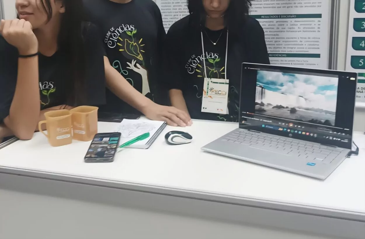 A imagem mostra que um grupo de estudantes está em uma bancada de apresentação, provavelmente em uma feira ou mostra científica. Eles vestem camisetas pretas com a inscrição “Clube de Ciências”. Sobre a mesa há diversos materiais: duas canecas, um celular, um caderno com anotações, uma caneta e um laptop aberto exibindo um vídeo de uma paisagem com água (parece uma cachoeira). Um dos alunos usa um crachá de evento.