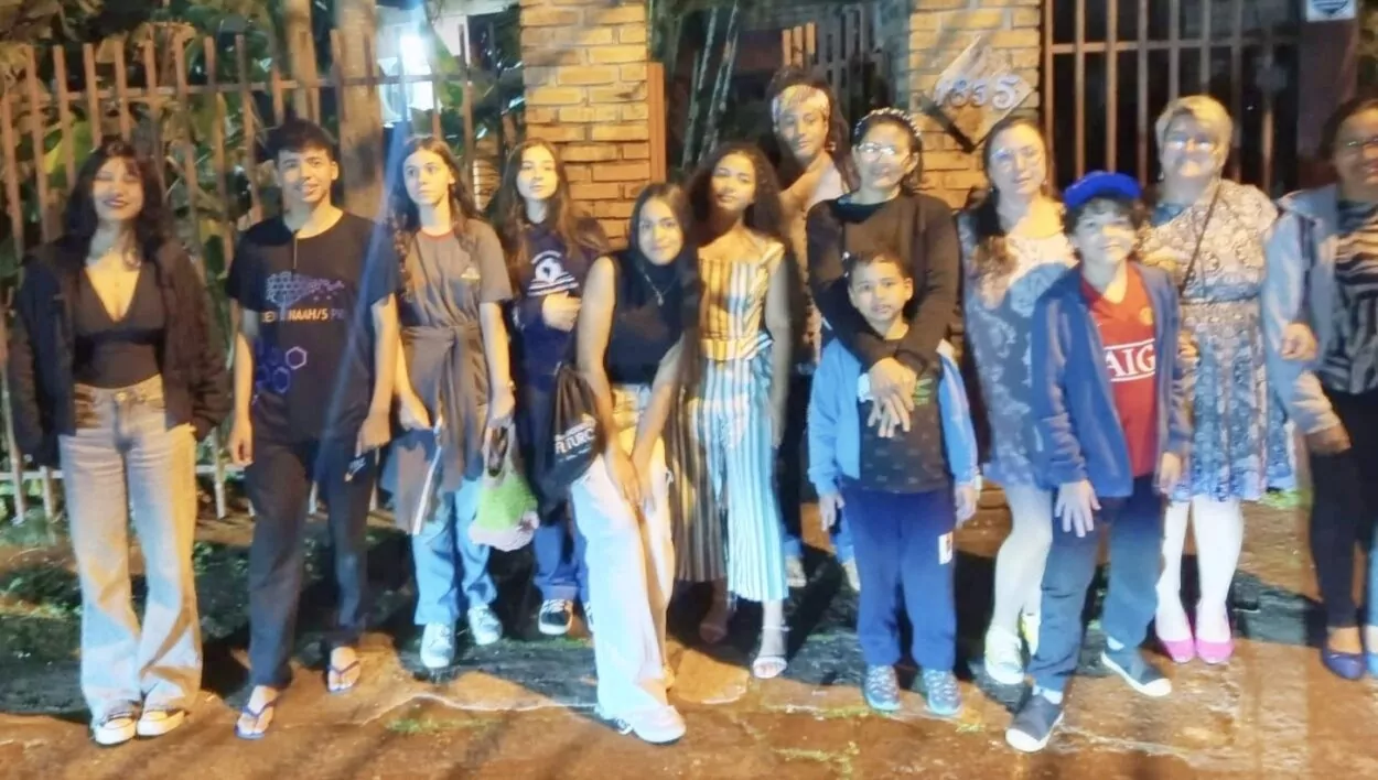 A imagem mostra um grupo grande de pessoas — incluindo adolescentes, crianças e alguns adultos, que estão reunidos à noite em frente a um portão com pilares de tijolos. O ambiente parece residencial ou institucional, com árvores e vegetação ao fundo. Todos estão posando para a foto, lado a lado, alguns sorrindo e outros com expressões mais neutras. As roupas são variadas, desde casuais até um pouco mais arrumadas, sugerindo um encontro ou atividade especial fora do horário escolar.
