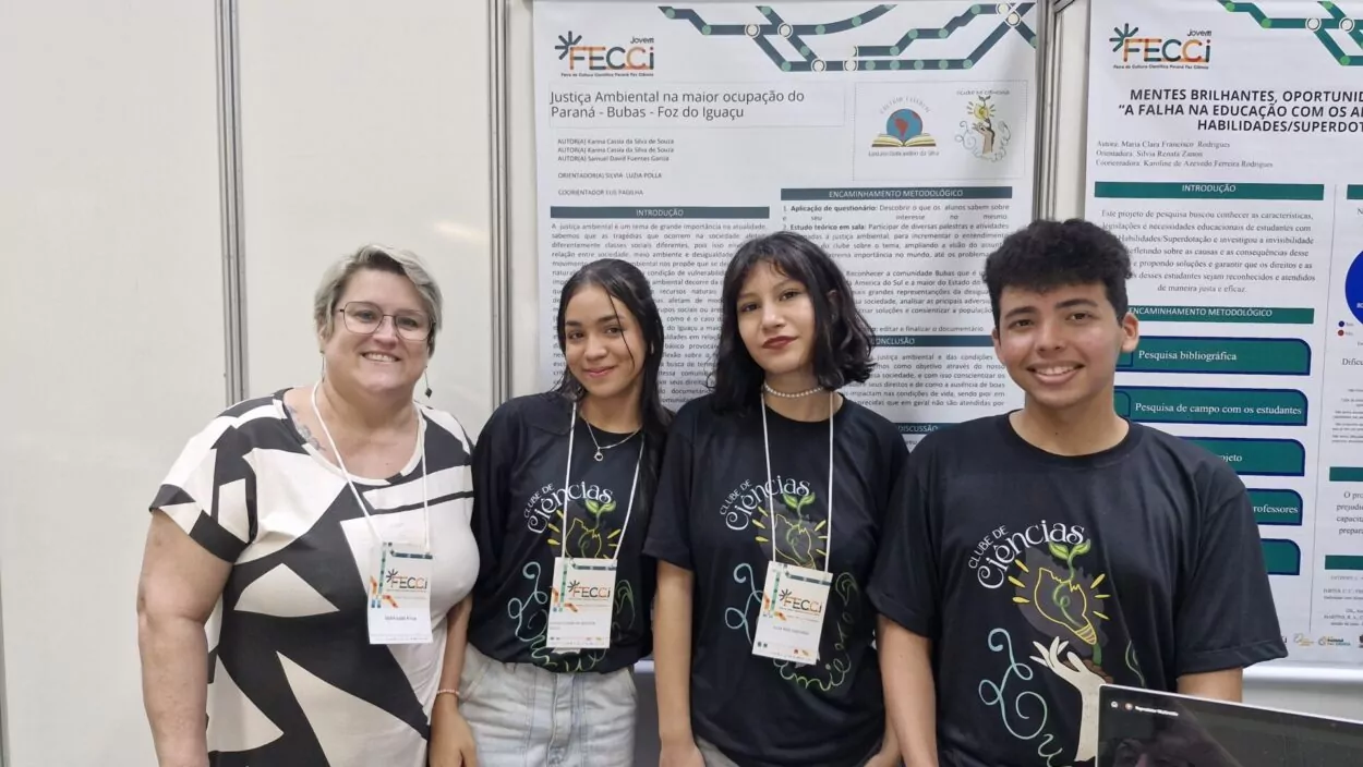 A imagem mostra que um grupo de estudantes e uma professora estão em uma bancada de apresentação, provavelmente em uma feira ou mostra científica. Os alunos vestem camisetas pretas com a inscrição “Clube de Ciências”. Todos posam para a foto. Atrás, há cartazes com a temática do projeto apresentado.