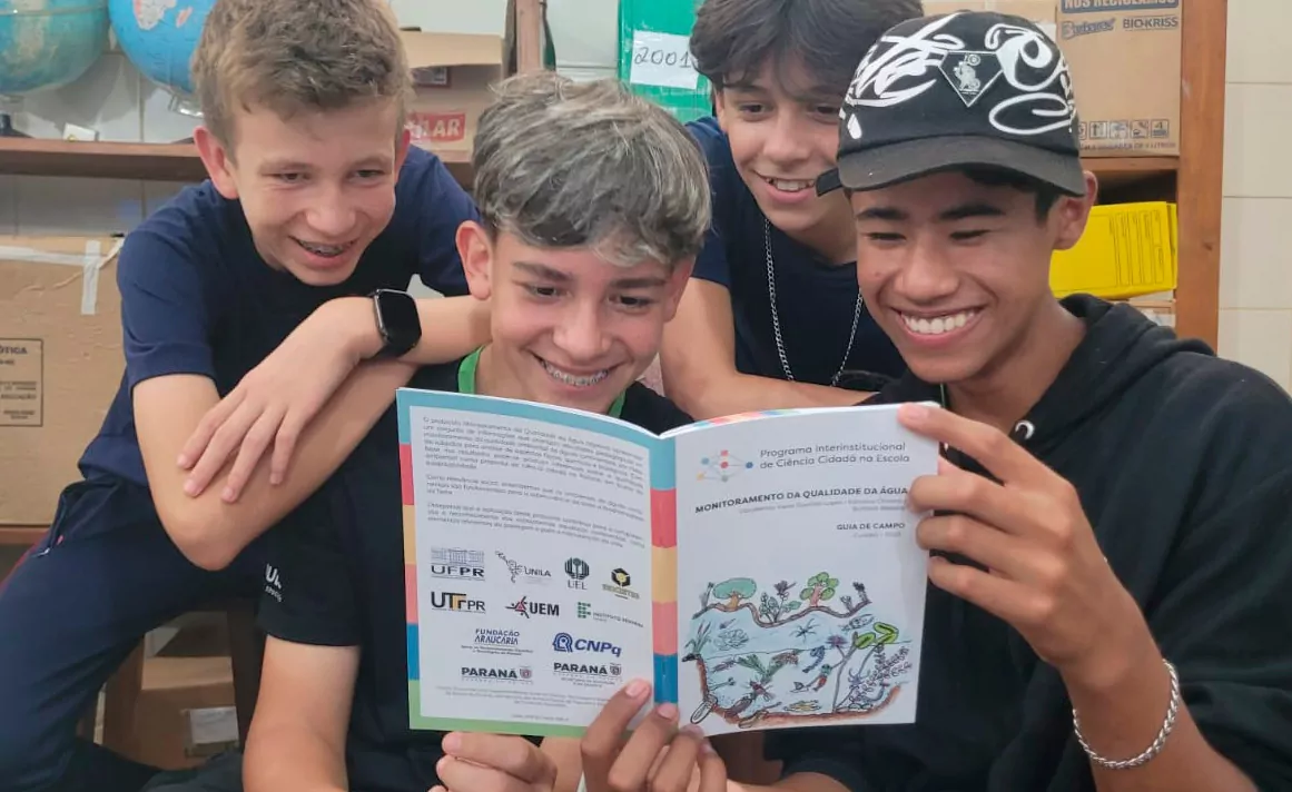 Foto em ambiente escolar ou laboratório. Quatro adolescentes estão reunidos bem próximos, sorrindo e olhando para um livreto aberto que seguram juntos. Dois estão sentados à frente e dois se inclinam por trás, apoiando os braços nos colegas. O livreto tem capa colorida e ilustrações de elementos naturais, como água, plantas e pequenos animais. No título, lê-se “Monitoramento da Qualidade da Água – Guia de Campo”, com logotipos de instituições acadêmicas e de pesquisa na parte inferior. Os jovens vestem roupas casuais, principalmente camisetas escuras; um deles usa boné preto com estampa branca. Ao fundo, há caixas, prateleiras e um globo terrestre, sugerindo um espaço educativo. A expressão geral do grupo é de interesse e entusiasmo pela leitura e pelo tema científico.